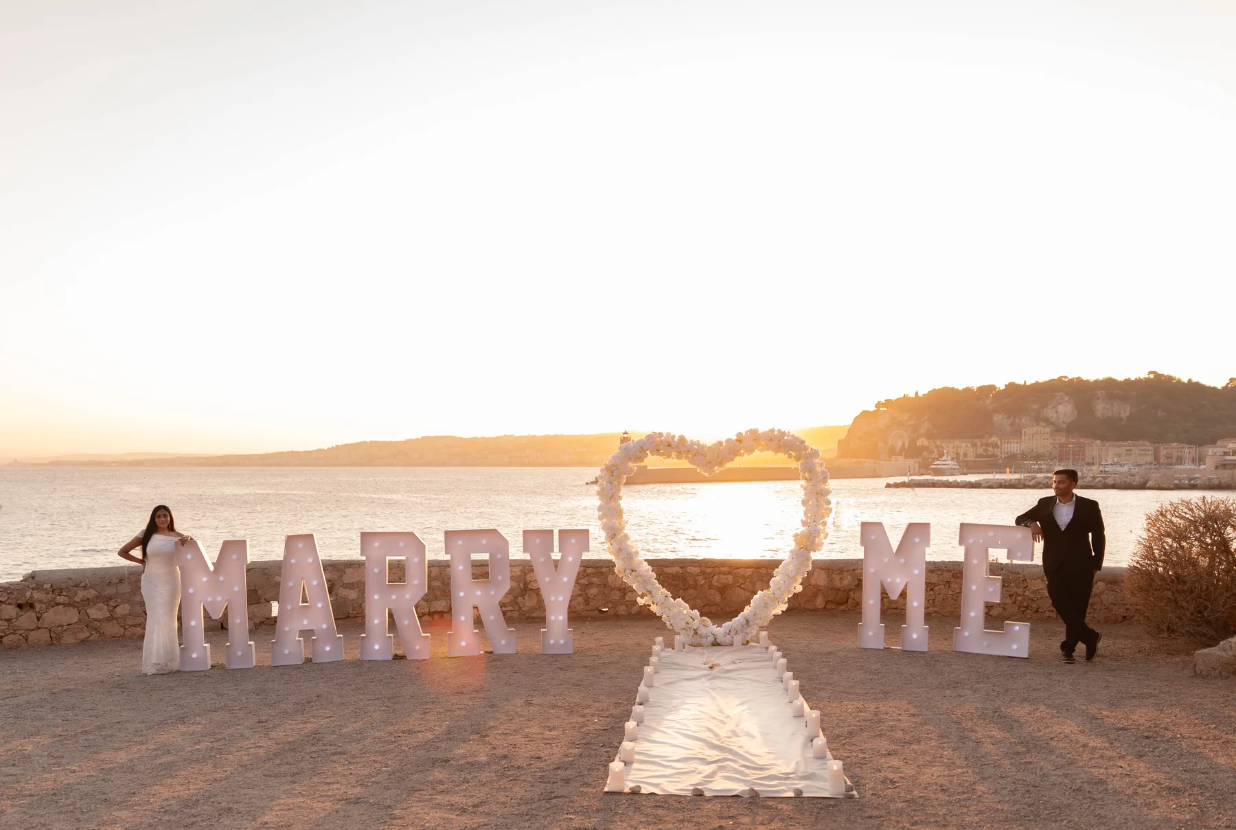 A &ldquo;Marry Me&rdquo; setup with a floral heart above the bay in Nice at sunset